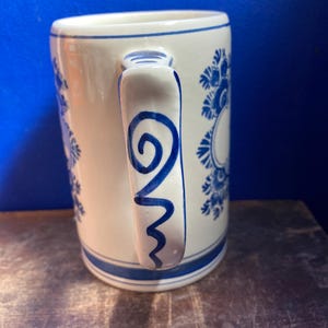May include: White ceramic mug with blue floral patterns and a blue handle design. The mug has a blue stripe at the base and rim. The handle features a blue swirl and zig-zag design.
