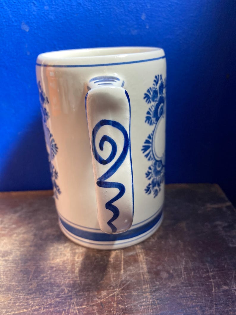 May include: White ceramic mug with blue floral patterns and a blue handle design. The mug has a blue stripe at the base and rim. The handle features a blue swirl and zig-zag design.