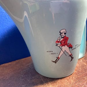 May include: A light blue ceramic pitcher with a curved handle. The pitcher features a graphic of a man in a red coat and white pants, with the text "BORN 1820 - STILL GOING STRONG".