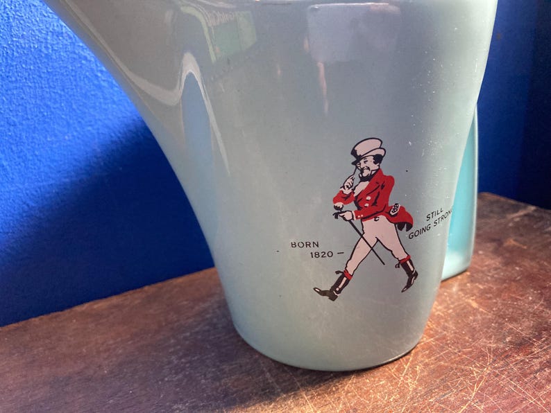 May include: A light blue ceramic pitcher with a curved handle. The pitcher features a graphic of a man in a red coat and white pants, with the text "BORN 1820 - STILL GOING STRONG".
