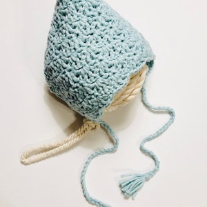 May include: A light blue crocheted baby hat with a white crocheted tie. The hat has a pointed top and two tassels.