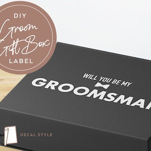 May include: A black gift box with a white decal that says "Will you be my groomsman" with a bowtie icon. The box is on a wooden surface. The image also includes a brown circle with the text "DIY Groom Gift Box Label" in white.