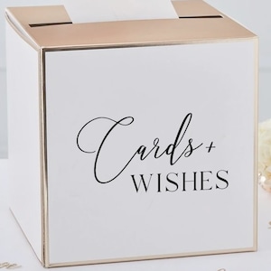 May include: White cardboard box with a gold trim and a slot on the top. The box has the text "Cards + Wishes" printed on the front in black cursive.