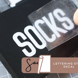 May include: A white vinyl sticker with the word "Socks" in a bold, blocky font. The sticker is being applied to a black surface. The sticker is labeled "Lettering Style Decal".