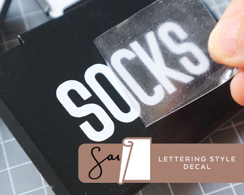 May include: A white vinyl sticker with the word "Socks" in a bold, blocky font. The sticker is being applied to a black surface. The sticker is labeled "Lettering Style Decal".