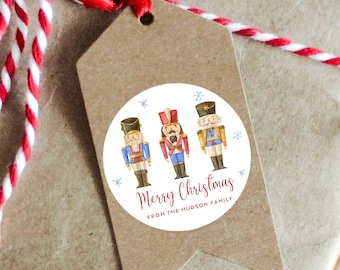 Könnte beinhalten: Runder weißer Aufkleber mit drei Nussknackerfiguren in Rot, Blau und Gold. Der Aufkleber hat ein rotes und weißes Schneeflockenmuster und den Text "Frohe Weihnachten von der Familie Hudson".