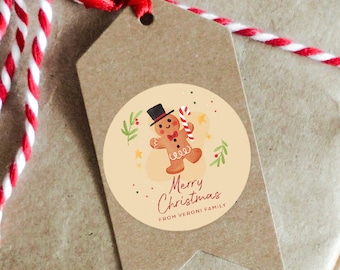 Könnte beinhalten: Ein runder, cremefarbener Geschenkanhänger mit einer Illustration eines Lebkuchenmanns. Der Lebkuchenmann trägt einen schwarzen Zylinder und hält einen Zuckerstock. Der Text "Merry Christmas" und "From Veroni Family" ist in Rot auf dem Anhänger gedruckt.