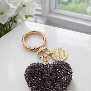 May include: A gold-toned keychain with a heart-shaped charm covered in dark brown crystals. The keychain includes a round clasp and a small circular tag. The tag reads "BRANDED GOODS & SUPPLIES SINCE 1982" and "NO40409".