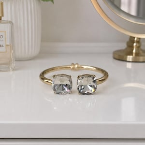 May include: A gold-toned cuff bracelet with two large, square, faceted gray crystals. The bracelet is displayed on a white surface, with a perfume bottle, a vase of white flowers, and a gold-framed mirror in the background.