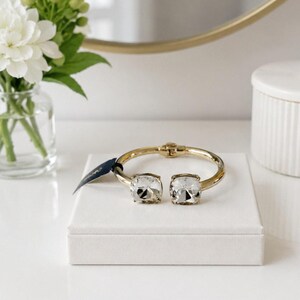 May include: A gold-toned hinged cuff bracelet featuring two large, square-cut clear crystals at the open ends. The bracelet has a polished metallic finish and includes a small dark blue tag attached to one side near the crystal setting.
