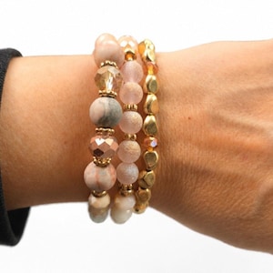 May include: Three beaded bracelets in shades of pink, gold, and gray. The bracelets feature a variety of bead shapes and sizes, including round, faceted, and irregular. The beads are strung on elastic cord, creating a stackable look.