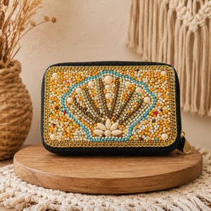 May include: A rectangular black zippered pouch featuring an intricate beaded design on the front. The beadwork depicts a stylized seashell motif in shades of yellow, cream, and turquoise, accented with gold metallic trim and small white pearls.