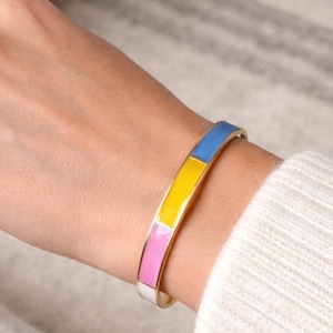 May include: Gold-tone bracelet with a colourful enamel design. The bracelet features a rectangular pattern of blue, yellow, and pink enamel. The bracelet is worn on a wrist, with a white sweater in the background.