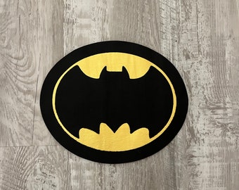Könnte beinhalten: Ein runder schwarzer Untersetzer mit einem goldenen Batman-Logo in der Mitte.