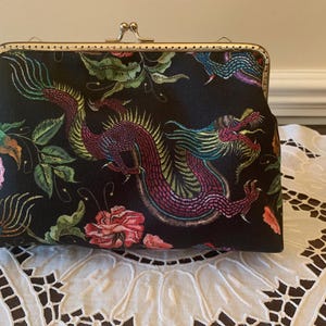 Puede incluir: Una cartera de mano de tela negra con un marco de metal dorado. La cartera presenta un diseño bordado colorido de un dragón y motivos florales.