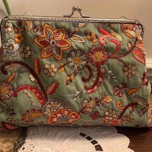 Puede incluir: Bolso de mano verde salvia con intrincados bordados florales en tonos rojos, naranjas y dorados. El bolso presenta un cierre de boquilla y una textura acolchada. El diseño floral incluye lentejuelas, añadiendo un toque de brillo.