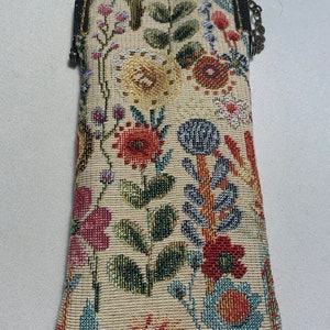 Op de afbeelding: Een beige clutch van bloemen-tapijt met een metalen sluiting en ketting. De clutch heeft een kleurrijk patroon van bloemen en bladeren in rood, blauw, geel en groen. De sluiting is donker bronskleurig.
