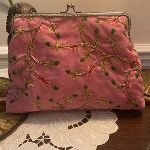 May include: Pink velvet clutch with a silver clasp. The clutch is adorned with gold and green embroidered floral patterns. The bag is resting on a white lace doily.