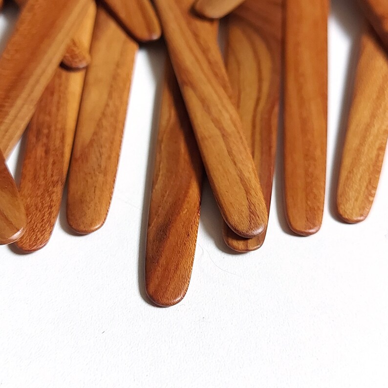 May include: A close-up of a pile of wooden butter knives. The knives are all made of light brown wood and have a smooth, rounded finish.