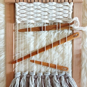May include: A wooden loom with a woven tapestry in white yarn. The loom has a fringe of grey yarn at the bottom. There are three wooden weaving tools on the loom.