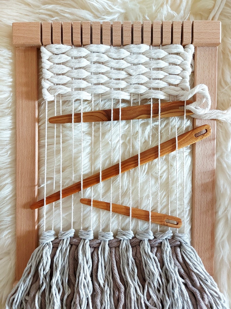 May include: A wooden loom with a woven tapestry in white yarn. The loom has a fringe of grey yarn at the bottom. There are three wooden weaving tools on the loom.