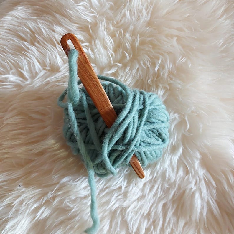 May include: A wooden yarn needle with a teal ball of yarn. The yarn needle is made of wood and has a smooth finish. The yarn is soft and fluffy.