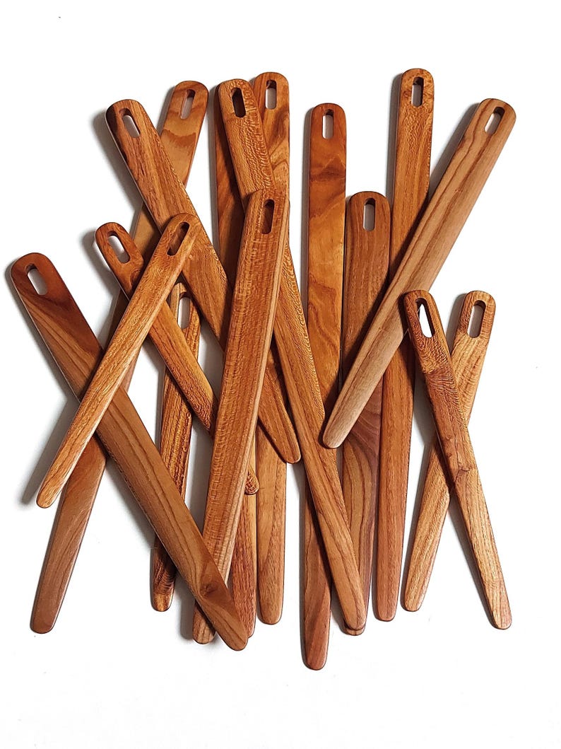 May include: A collection of wooden knitting needles, all in various shades of brown, are stacked on top of each other. The needles are all the same size and shape, and they have a smooth, polished finish.