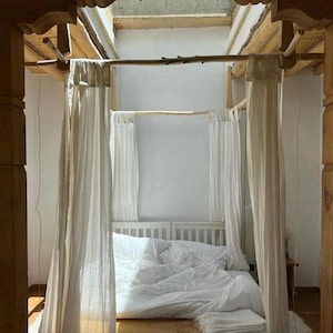 May include: A white canopy bed with white curtains and a white duvet. The bed is made of wood and has a wooden frame. The bed is in a room with a wooden ceiling and a skylight.