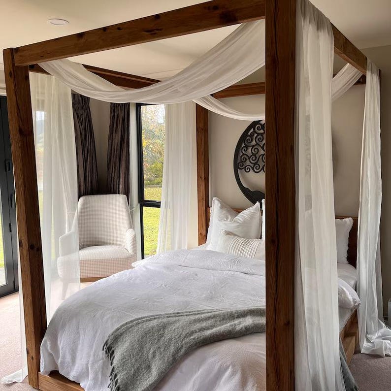 May include: A wooden four-poster bed frame with white sheer curtains and a white duvet and gray throw blanket. The bed has a black metal tree of life wall decor above the headboard.