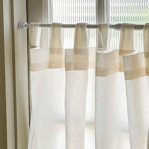 May include: White linen curtains with a beige band at the top, hanging from a white curtain rod. The curtains are semi-sheer, allowing light to filter through. The window is visible in the background.