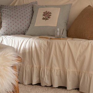 May include: A white couch with a ruffled skirt, three pillows, and a glass on a book. The pillows are patterned with blue and white gingham, small pink flowers, and brown burlap.