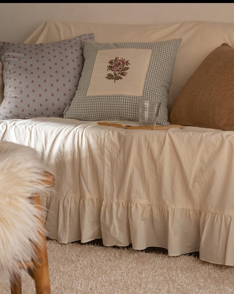 May include: A white couch with a ruffled skirt, three pillows, and a glass on a book. The pillows are patterned with blue and white gingham, small pink flowers, and brown burlap.