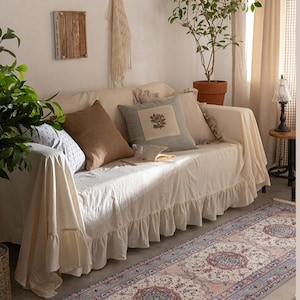 May include: A white sofa with a frilled skirt and cushions sits in a living room with a patterned rug on the floor. The rug has a light blue background with a pink and white floral design.