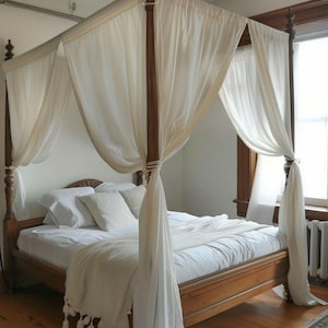 May include: A wooden four-poster bed with white sheer curtains draped over the top. The bed is made with white linens and a white throw blanket with tassels.