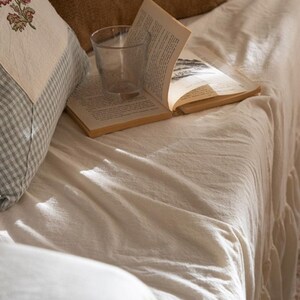 May include: A white linen throw blanket with a glass of water and a book on top. The blanket is on a couch with a pillow with a floral pattern.