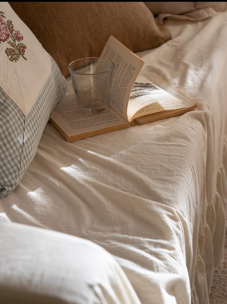 May include: A white linen throw blanket with a glass of water and a book on top. The blanket is on a couch with a pillow with a floral pattern.