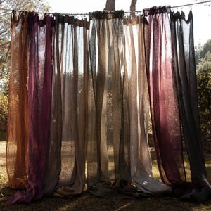 May include: Sheer, flowing curtains in shades of brown, purple, and tan hang outdoors. The fabric is lightweight and appears to be linen or a similar material. The curtains are suspended from a rod, creating a natural, airy aesthetic.