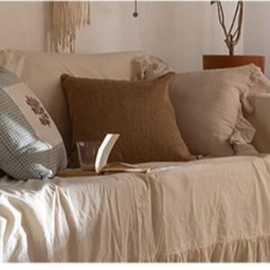 May include: A white couch with a ruffled edge, covered with a white throw blanket. The couch has several pillows, including a brown pillow, a white pillow, and a patterned pillow. A lamp with a clear glass shade sits on a wooden table in front of the couch.