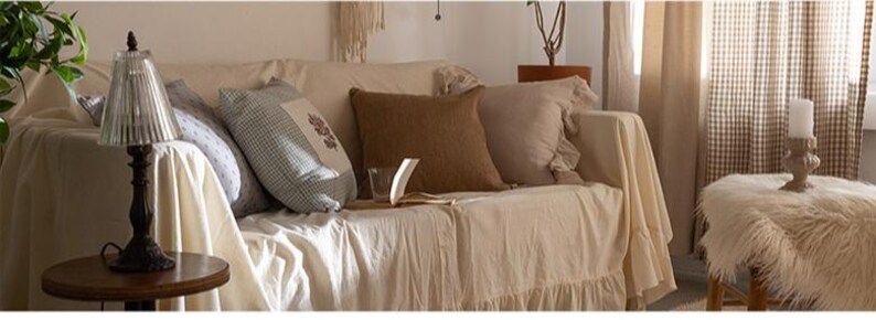 May include: A white couch with a ruffled edge, covered with a white throw blanket. The couch has several pillows, including a brown pillow, a white pillow, and a patterned pillow. A lamp with a clear glass shade sits on a wooden table in front of the couch.