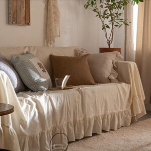 May include: A white couch with a ruffled skirt and a variety of throw pillows, including a brown pillow, a blue and white checkered pillow, and a white pillow with a floral design. The couch is in a living room with a lamp, a plant, and a window.