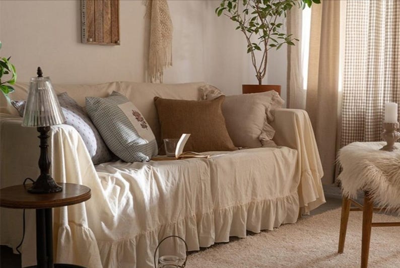 May include: A white couch with a ruffled skirt and a variety of throw pillows, including a brown pillow, a blue and white checkered pillow, and a white pillow with a floral design. The couch is in a living room with a lamp, a plant, and a window.