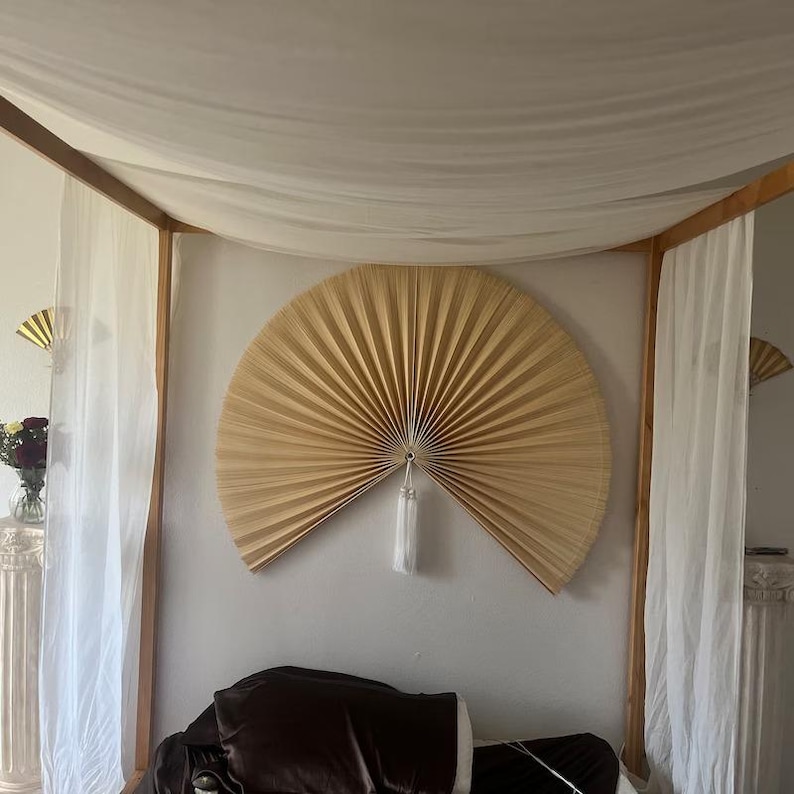 May include: A large, light brown, woven fan with a white tassel hanging from the center. The fan is mounted on a white wall in a bedroom with white curtains and a bed with a brown headboard.