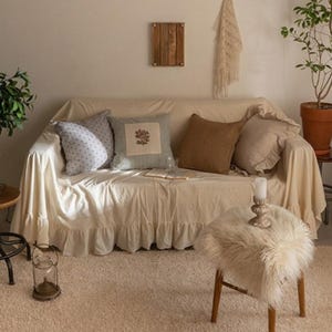 May include: A white couch with a ruffled skirt, covered with a white throw blanket and several throw pillows. The pillows are patterned with blue and white floral designs, brown fabric, and a light blue and white striped design. The couch is in a living room setting with a rug, a small table with a candle, and a furry stool.
