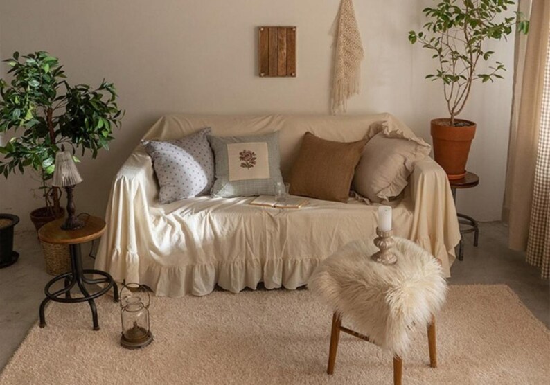 May include: A white couch with a ruffled skirt, covered with a white throw blanket and several throw pillows. The pillows are patterned with blue and white floral designs, brown fabric, and a light blue and white striped design. The couch is in a living room setting with a rug, a small table with a candle, and a furry stool.