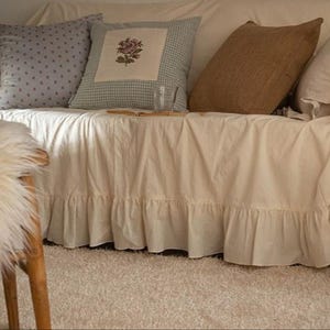 May include: A white couch with a ruffled skirt, featuring a variety of throw pillows in different patterns and textures. The couch is decorated with a floral pillow, a brown pillow, and a gray polka dot pillow. The couch is sitting on a light beige rug.