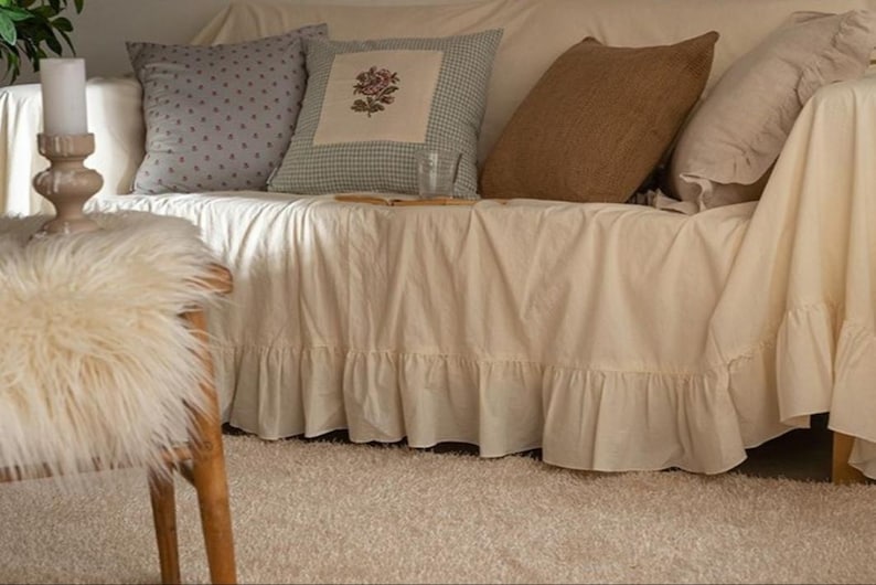 May include: A white couch with a ruffled skirt, featuring a variety of throw pillows in different patterns and textures. The couch is decorated with a floral pillow, a brown pillow, and a gray polka dot pillow. The couch is sitting on a light beige rug.