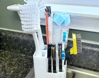 May include: A white plastic organizer holding various cleaning tools, including two white dish brushes, a coral and black brush, a bottle brush with blue sponges, three small black-handled detail brushes, and a yellow-handled wire brush.