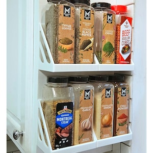 May include: A white, two-tiered spice rack attached to a cabinet door, filled with various spice jars. The jars contain thyme leaves, coarse black pepper, chopped chives, beef bouillon cubes, Montreal steak seasoning, granulated garlic, onion, and chili powder.