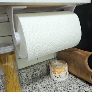 May include: White paper towel holder mounted under a cabinet with a roll of white paper towels. The holder is made of white plastic and has a simple design.