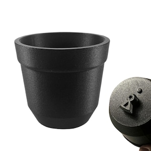May include: A black, textured, cylindrical container with a wide opening, resembling a small flower pot. A matching black lid with a raised design is held nearby. The items appear to be made of a matte material.